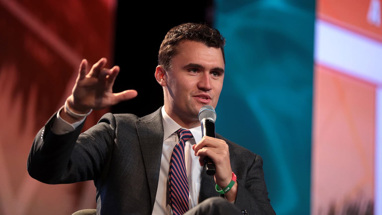 Charlie Kirk seated on stage, speaking into a microphone